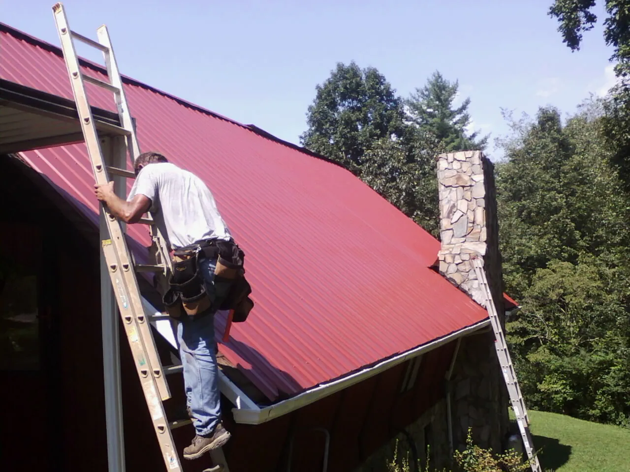 Roofer on a ladder beside a red metal roof and stone chimney — Soffit & Fascia Repair by Roofing Brothers in Oak Brook